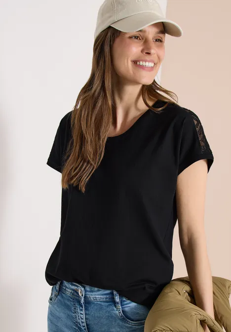 Eine Frau trägt ein schwarzes T-Shirt, blaue Jeans und eine Cap, hält eine olivgrüne Jacke.