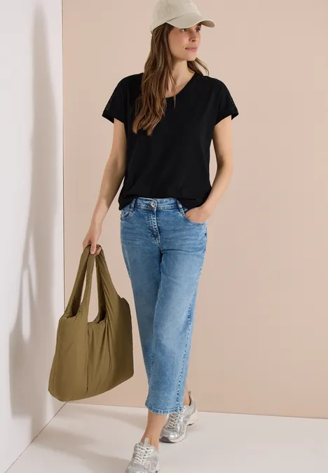 Frau im schwarzen T-Shirt, blauer Jeanshose, silbernen Schuhen und breitem Hut trägt eine olivgrüne Tasche.