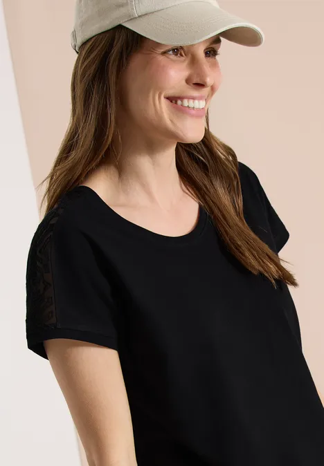 Eine Frau mit schwarzem T-Shirt und weißer Cap vor hellem Hintergrund, Gesicht unkenntlich.