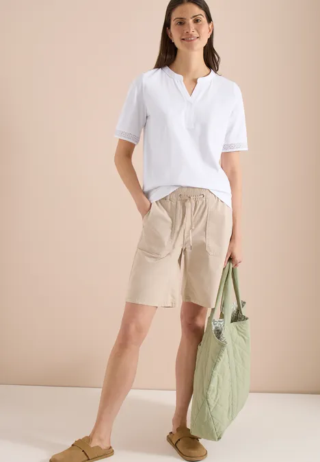 Frau im weißen T-Shirt, beigen Bermudashorts, beigen Sandalen mit grüner Tasche.