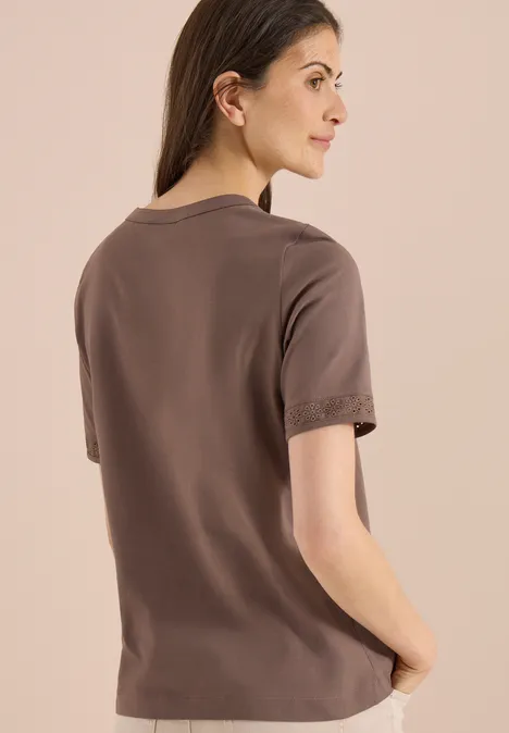 Frau in einfachem braunen T-Shirt, von hinten fotografiert vor hellem Hintergrund.