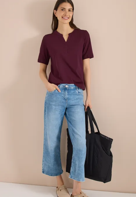 Vrouw met een bordeauxrood t-shirt, wijde cropped jeans en een zwarte tas tegen een beige achtergrond.