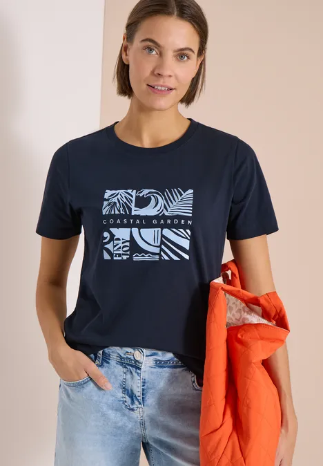 Shirt met ronde hals en frontprint universal blue Vrouw draagt marineblauw T-shirt met grafische print en houdt een oranje jas vast.