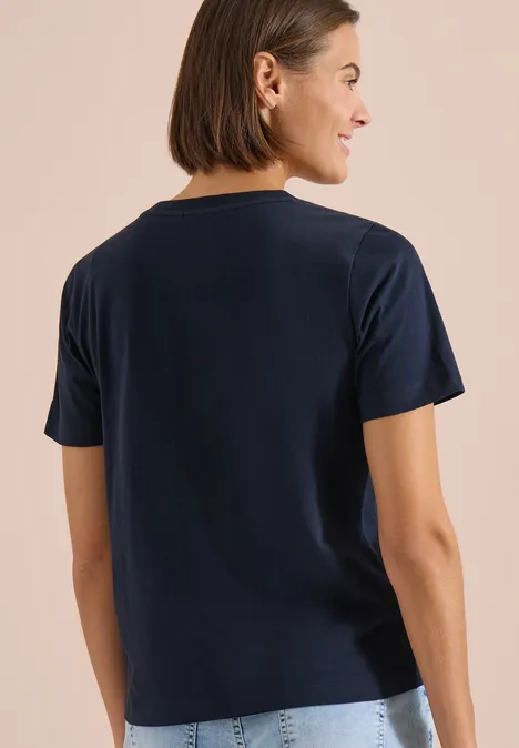 Shirt met ronde hals en frontprint universal blue Rugaanzicht van een persoon in een donkerblauw T-shirt en lichte broek tegen een beige achtergrond.