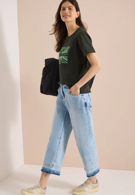 Shirt met ronde hals en frontprint raw khaki Vrouw in profiel in een minimalistisch studio, draagt een zwart t-shirt met groene print, wijde lichtblauwe jeans en een grote tas.