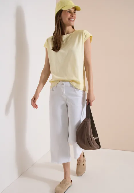 Frau in gelbem T-Shirt, weißer Culotte, gelber Mütze und brauner Tasche vor neutralem Hintergrund.
