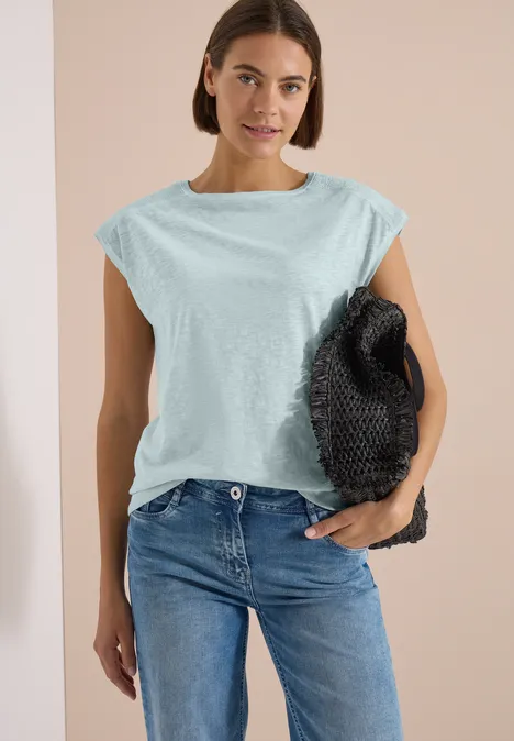Frau trägt hellblaues ärmelloses Oberteil, blaue Jeans und hält schwarze Tasche.