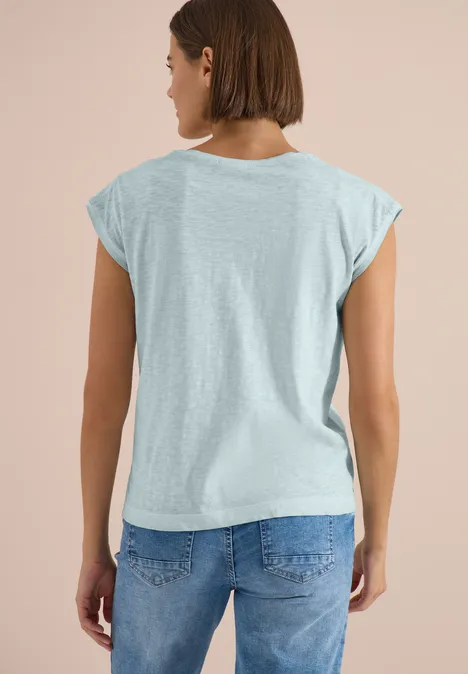 Eine Frau von hinten in hellgrauem T-Shirt und blauer Jeans vor hellem Hintergrund.
