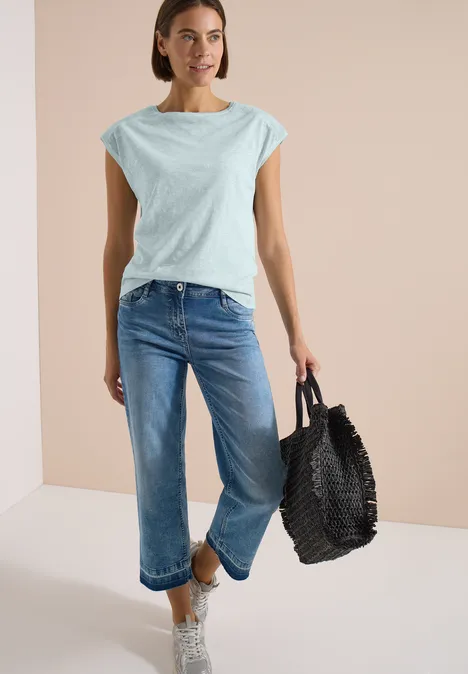 Frau mit hellblauem T-Shirt, blauen Jeans und schwarzer geflochtener Tasche vor neutralem Hintergrund.