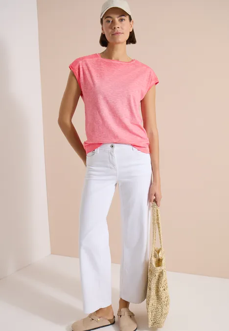 T-Shirt aus Jersey im Garment Dye Look coral Frau trägt pinkes T-Shirt, weiße gerade Hose und Basttasche vor hellem Hintergrund.