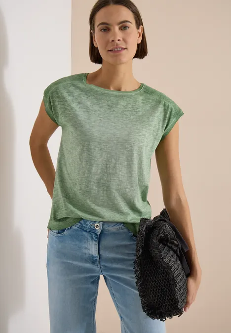 T-shirt en jersey au look garment dye deep matcha green Eine stehende Person in grüner Bluse und Jeans hält eine schwarze Tasche.