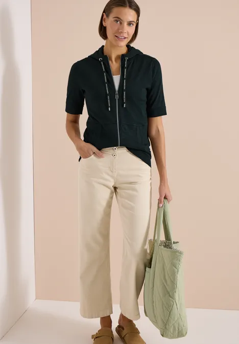 Frau in schwarzem Kurzarm-Zipshirt, hellem Culotte-Hose und gestepptem grünen Shopper