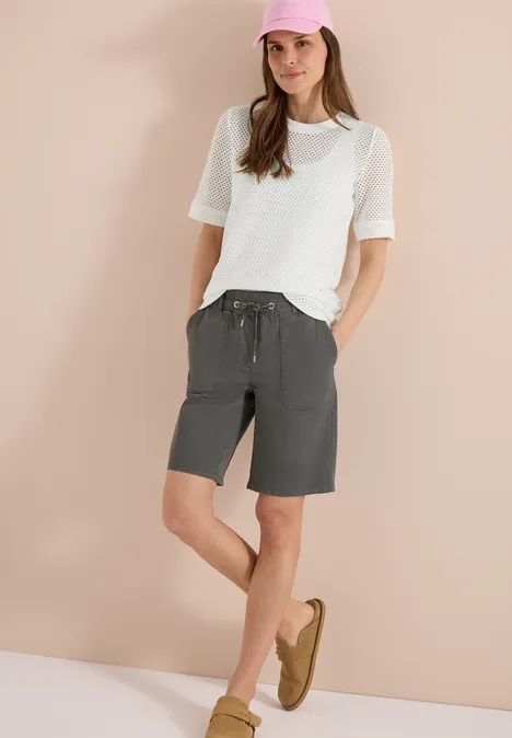 Shirt met ronde hals in ajour look vanilla white Vrouw staat voor een beige achtergrond, draagt wit T-shirt, grijze bermudashort en bruine loafers.