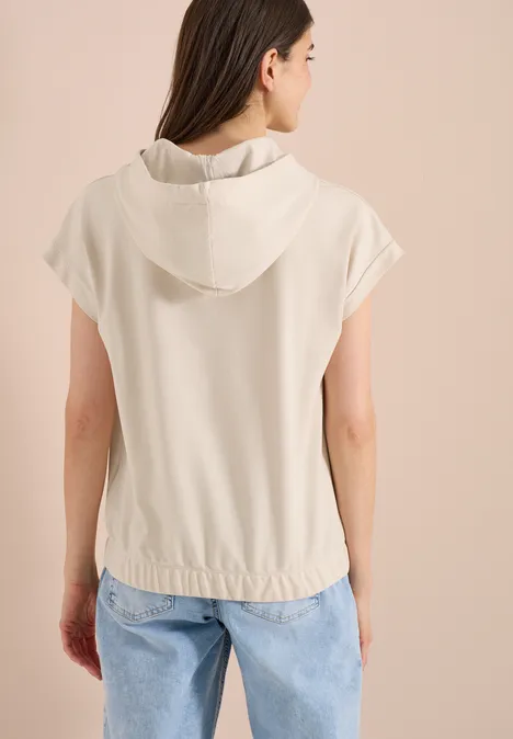 Vrouw van achteren in een beige capuchonshirt met korte mouwen en een losse pasvorm tegen een lichte achtergrond