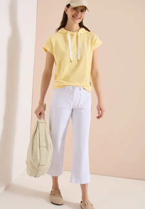 Vrouw in gele capuchon-top, witte flared broek, beige pet en loafers, met een doorgestikt tas.