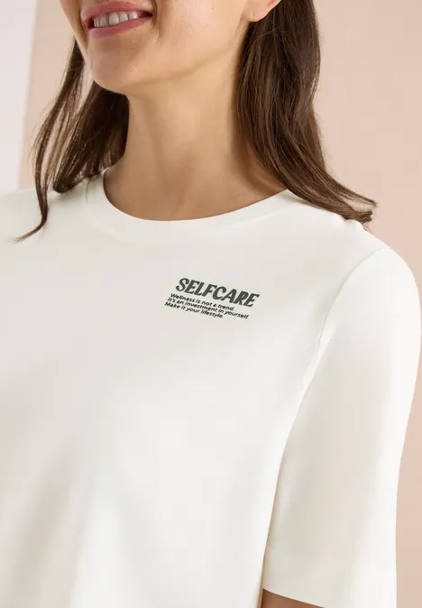 Eine Frau trägt ein weißes Shirt mit kleinem 'SELF-CARE'-Aufdruck.