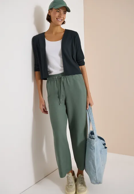 Een persoon staat voor een muur, draagt groene joggingbroek, wit T-shirt, donkerblauw vest, pet en lichtblauwe tas.