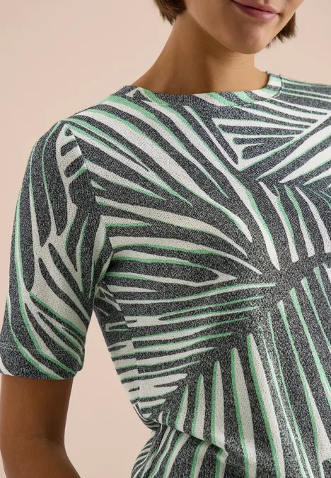 Shirt met boothals en print palm breeze green Close-up van een T-shirt met korte mouwen en groot bladpatroon in groen en zwart op een lichte achtergrond.
