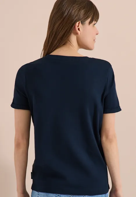 Frau von hinten mit dunkelblauem Kurzarmshirt und langen braunen Haaren vor beigem Hintergrund.