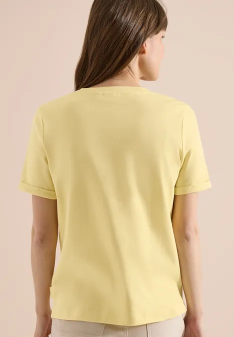 Eine Frau von hinten, die ein hellgelbes Kurzarmshirt trägt vor hellem Hintergrund.