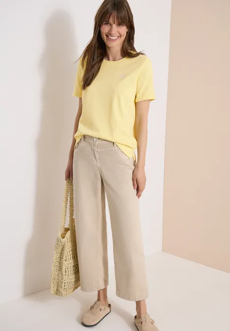 Frau vor hellem Hintergrund trägt gelbes T-Shirt, bequeme beige Hose, helle Schuhe und hält Netztasche.