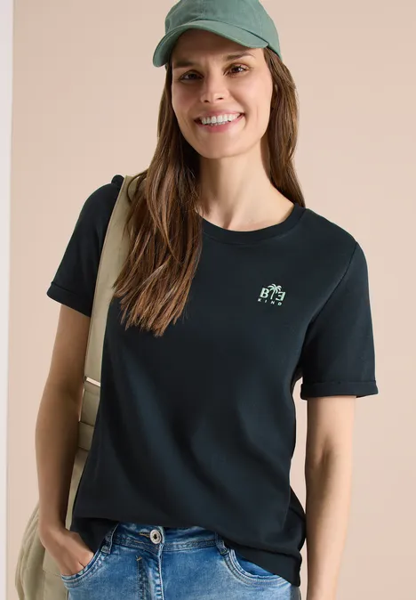 Vrouw in zwart t-shirt met klein logo, spijkerbroek, pet en schoudertas voor lichte achtergrond