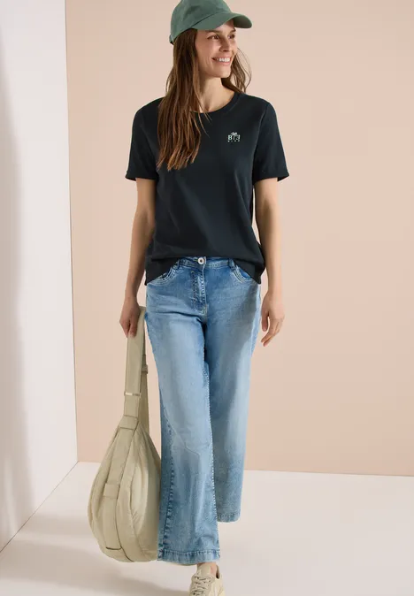 Vrouw in zwart t-shirt met walvisprint, blauwe jeans, witte sneakers, groene pet en een grote lichtgroene tas.