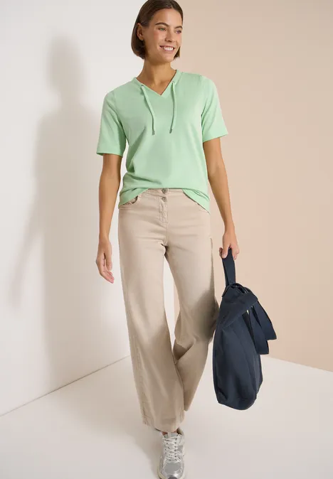 Gestreept shirt juicy lime Vrouw in groen capuchon-T-shirt, beige wijd uitlopende broek en blauwe tas bij lichte muur.