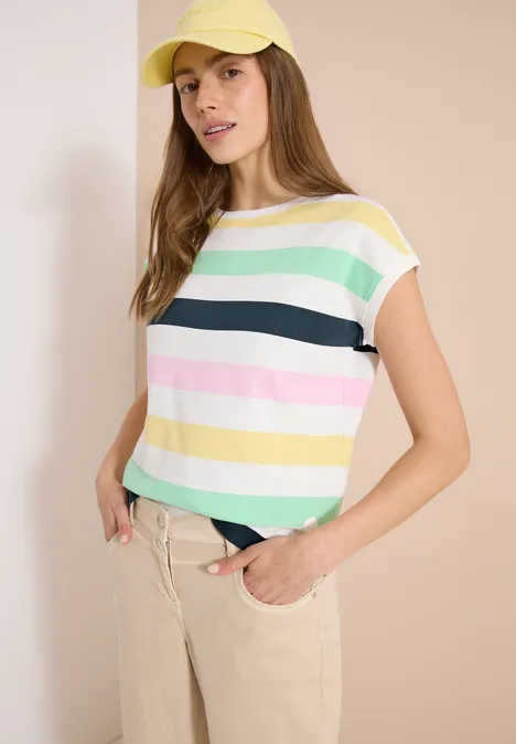 Frau in pastellfarben gestreiftem T-Shirt und beigen Hosen, stehend mit Händen in den Taschen vor beigem Hintergrund.