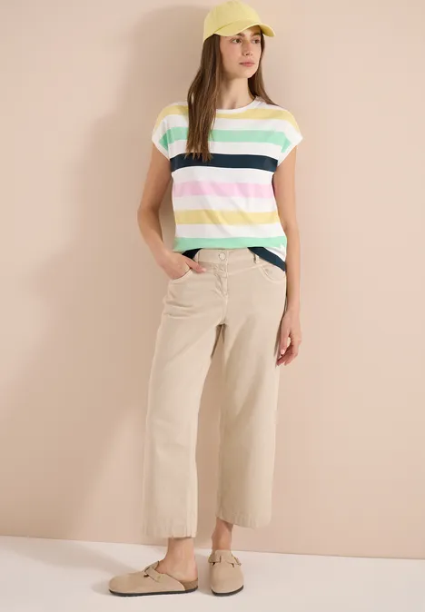 Eine Person trägt bunt gestreiftes T-Shirt, beige Culotte-Hosen und Slipper vor hellem Hintergrund.