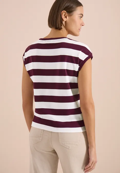 Eine Frau steht mit dem Rücken zur Kamera und trägt ein weiß-bordeauxgestreiftes T-Shirt mit beigen Hosen.