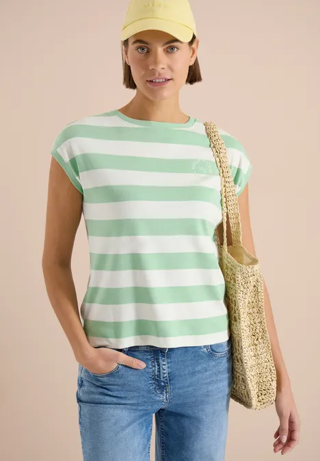 Frau trägt grün-weiß gestreiftes T-Shirt, Jeans und eine geflochtene Strohtasche über der Schulter.