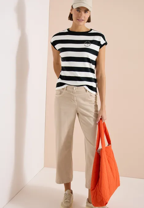 Frau mit schwarz-weiß gestreiftem Shirt, beigen Hosen, Sandalen und orangefarbener Tasche vor neutralem Hintergrund.