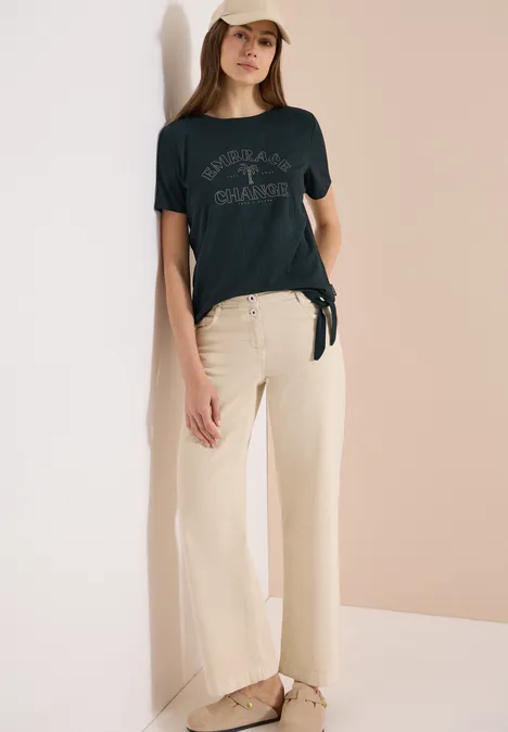 Frau trägt schwarzes T-Shirt mit Knoten, beige weite Hose und Sandalen vor hellem Hintergrund.