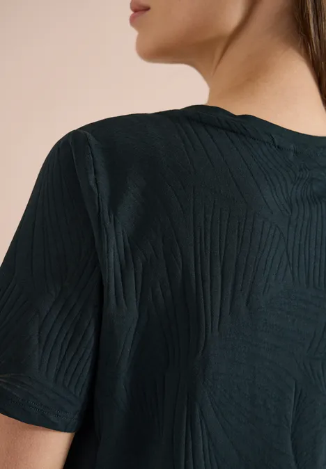 Rückansicht einer Frau in dunkelgrünem, texturiertem T-Shirt mit kurzem Arm.