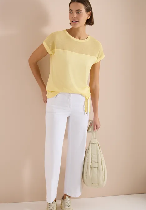 Model trägt transparentes gelbes Kurzarmshirt mit Knotensaum, weiße Cropped-Hose und pastellgrüne Tasche.