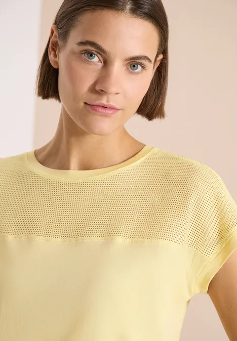 Frau in pastellgelbem, kurzärmeligem Stricktop vor hellem, neutralem Hintergrund.