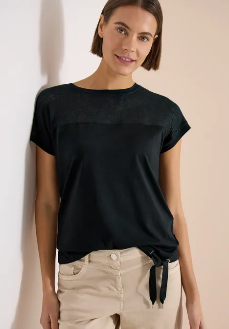 Vrouw leunt tegen muur, draagt zwart t-shirt met strik en lichte broek.