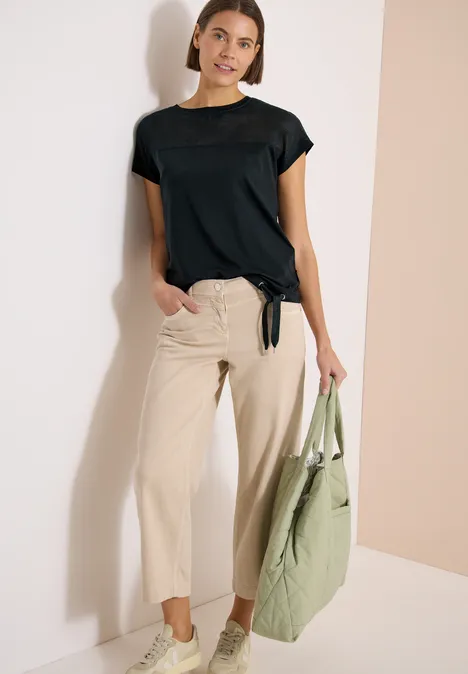 Model draagt een zwart t-shirt met korte mouwen, een beige broek en houdt een groene tas vast, leunend tegen een muur.