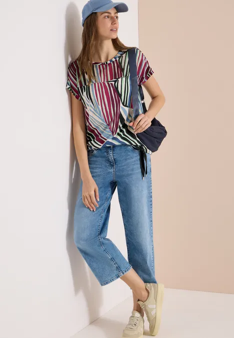 Femme adossée, chemisier rayé coloré, jean ample, casquette, sac bandoulière et baskets claires.