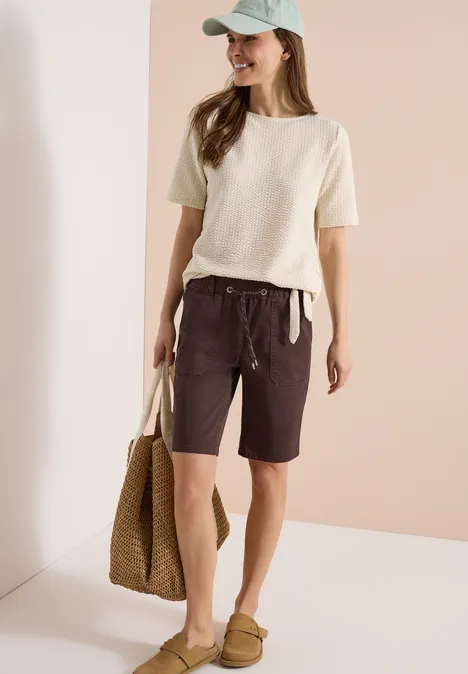 Shirt in seersucker stof pearl beige Vrouw in witte T-shirt, bruine short, groene pet, strooien tas en mocassins.