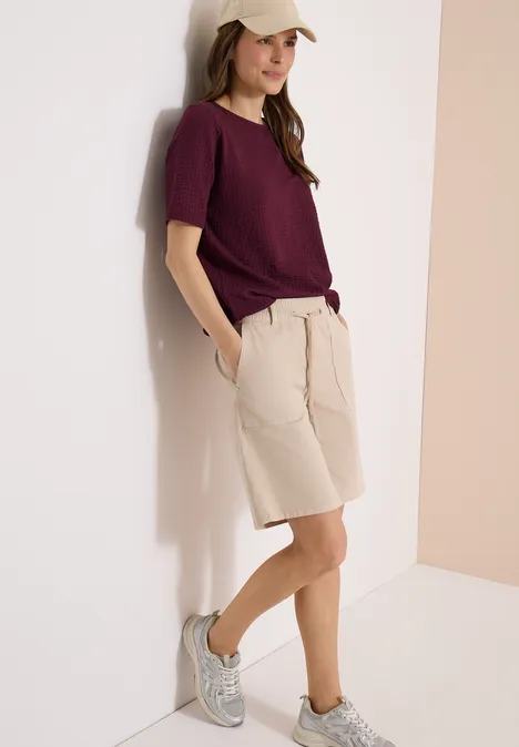 Shirt in seersucker stof sweet grape red Een persoon leunt tegen een muur, draagt een bordeauxrood t-shirt, beige shorts en grijze sneakers.