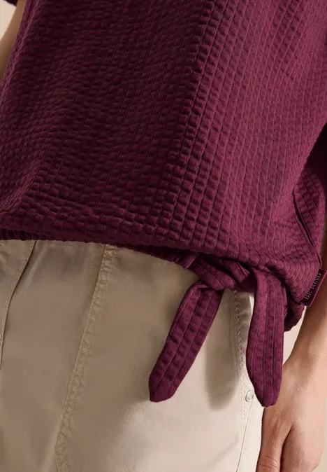 Shirt in seersucker stof sweet grape red Donkerpaarse gewatteerde top met zijstrik, gecombineerd met beige broek.