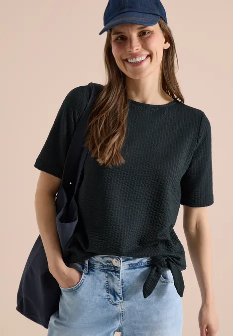 Shirt in seersucker stof palm breeze green Vrouw met geknoopt zwart kort shirt, blauwe spijkerbroek, pet en schoudertas tegen neutrale achtergrond.