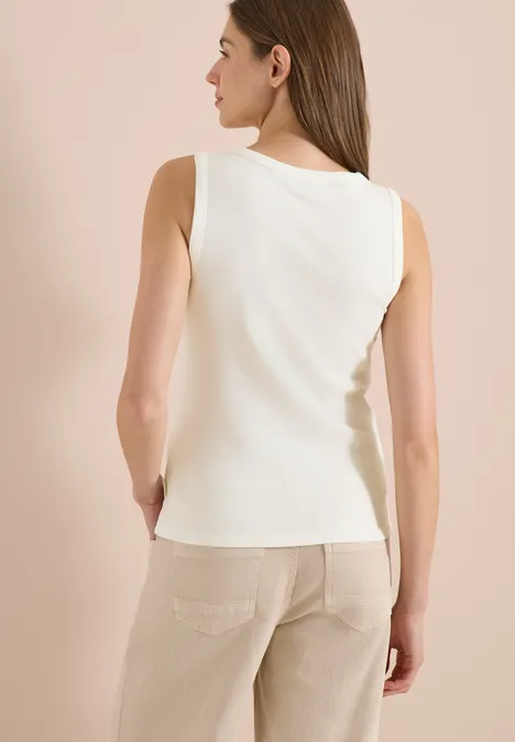 Vrouw van achteren in een witte mouwloze top en een beige broek tegen een neutrale achtergrond.