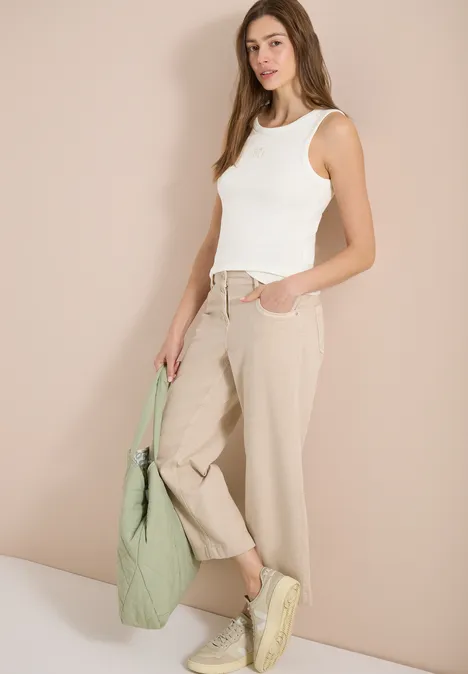 Vrouw met wit hemdje, beige broek, gele sneakers, houdt een groene tas vast.