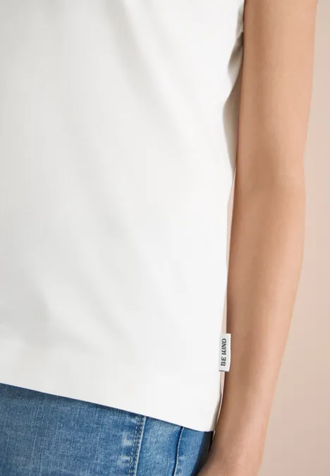 Weißes ärmelloses Oberteil aus leichtem Stoff, sitzt locker über blauen Jeans, mit seitlichem Label.