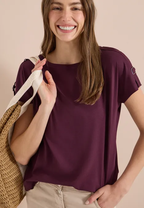 Frau in lila Kurzarmshirt und beiger Hose mit Strohtasche über der Schulter.