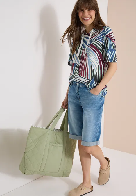 Hoodie shirt met print palm breeze green Vrouw in gestreept shirt, denim bermuda en comfortabele klompen, met een grote groene gewatteerde tas.