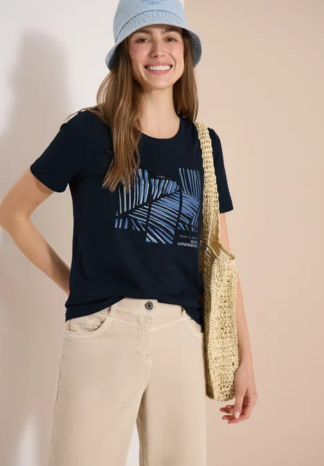 Frau in dunkelblauem T-Shirt mit Muster, hellen Hosen, Strohtasche und Hut vor weißem Hintergrund.